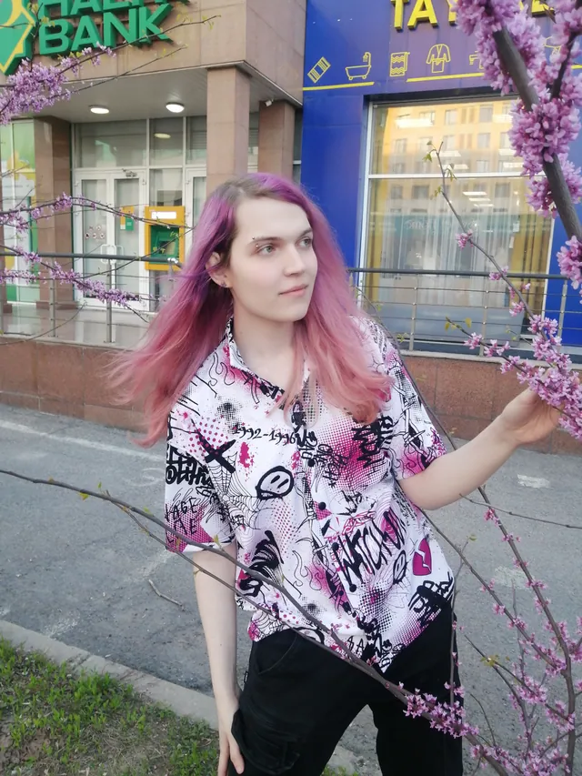 Liz standing next to a blooming tree in front of a bank, pink hair, patterned shirt and black trousers