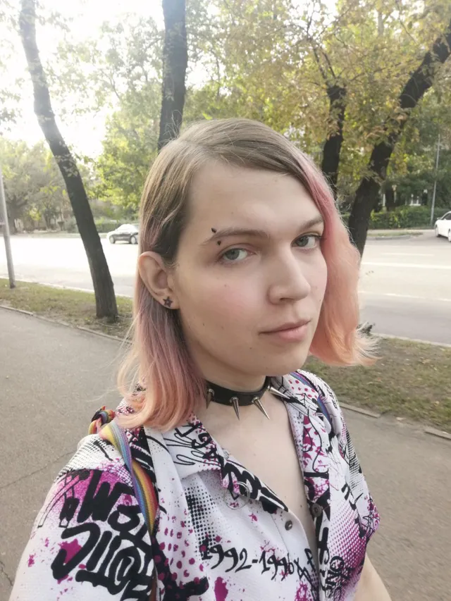 Liz on a tree-lined street, blonde hair fading to pink, looking at the camera