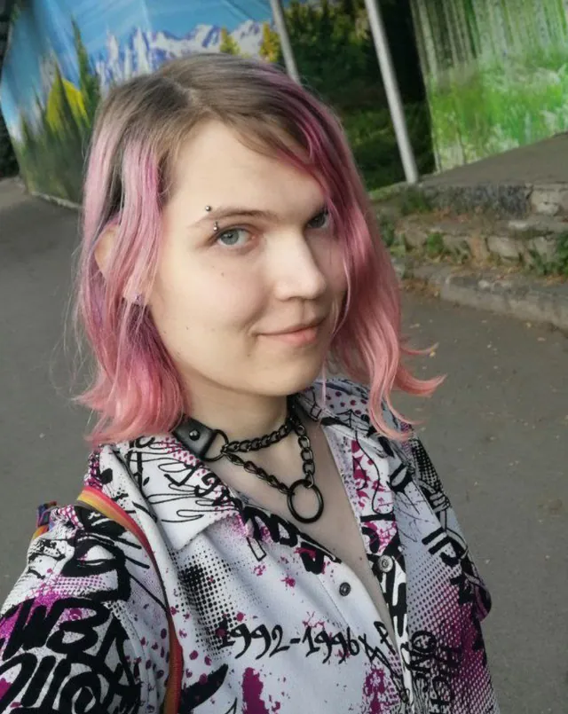 Liz with shoulder-length pink hair and a choker, in a black-and-white patterned shirt, smiling a little
