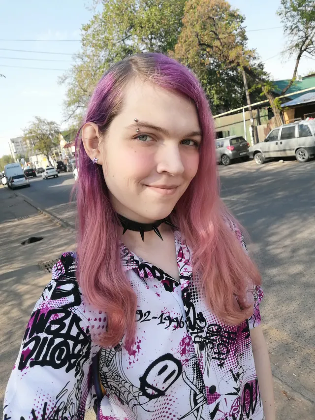 Liz on a sunny street with pink hair and a spiky choker, smirking